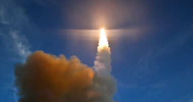 (Representative only.) A bright column of flame and smoke illuminates the sky as a missile is launched, ascending rapidly against a blue and white cloudy backdrop. Photo: Wikimedia Commons