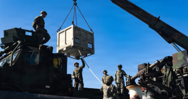 Service members use a crane to load a large supply crate onto a military vehicle during logistics operations.