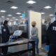 Two airport security officers monitor a passenger during screening beside an X-ray scanner and walk-through body scanner at a checkpoint.
