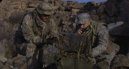 Two military personnel in camouflage uniforms operate a rugged field computer in a rocky outdoor environment, supporting on-site data processing and communications.