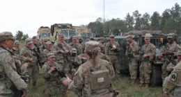 Group of U.S. soldiers in camouflage receiving a field briefing beside military vehicles during a training exercise.