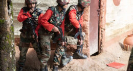 Three Indian Army soldiers wearing camouflage uniforms and helmets take cover beside a building during a cordon and search training exercise, holding rifles and communicating by radio.