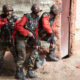 Three Indian Army soldiers wearing camouflage uniforms and helmets take cover beside a building during a cordon and search training exercise, holding rifles and communicating by radio.