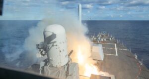 Missile launches vertically from the bow of a U.S. Navy destroyer, flames and smoke rising above the deck-mounted launch system in open ocean.
