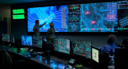 Military personnel analyze data on multiple screens in a command center with maps, analytics dashboards, and sensor feeds displayed.