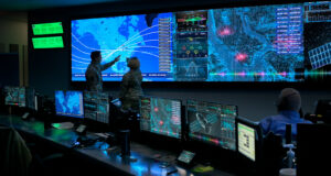 Military personnel analyze data on multiple screens in a command center with maps, analytics dashboards, and sensor feeds displayed.