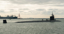 Virginia-class submarine surfaced at sea with crew on deck near a port facility under cloudy skies.