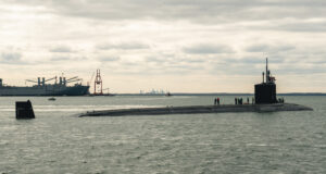 Virginia-class submarine surfaced at sea with crew on deck near a port facility under cloudy skies.