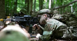 US Army National Guard soldier takes cover during infantry training exercise in wooded terrain.