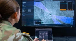 Soldier operating a laptop connected to a large screen displaying a battlefield map with targeting overlays and mission coordination interface.