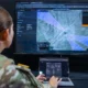 Soldier operating a laptop connected to a large screen displaying a battlefield map with targeting overlays and mission coordination interface.