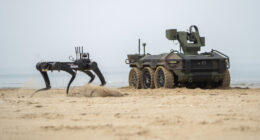 A quadruped military robot and Hyundai Rotem HR-Sherpa unmanned ground vehicle operate on sandy terrain.