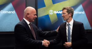Two executives shake hands in front of Saab and Comand AI displays during the announcement of a strategic partnership focused on AI-powered military planning and decision-support technologies.
