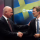 Two executives shake hands in front of Saab and Comand AI displays during the announcement of a strategic partnership focused on AI-powered military planning and decision-support technologies.